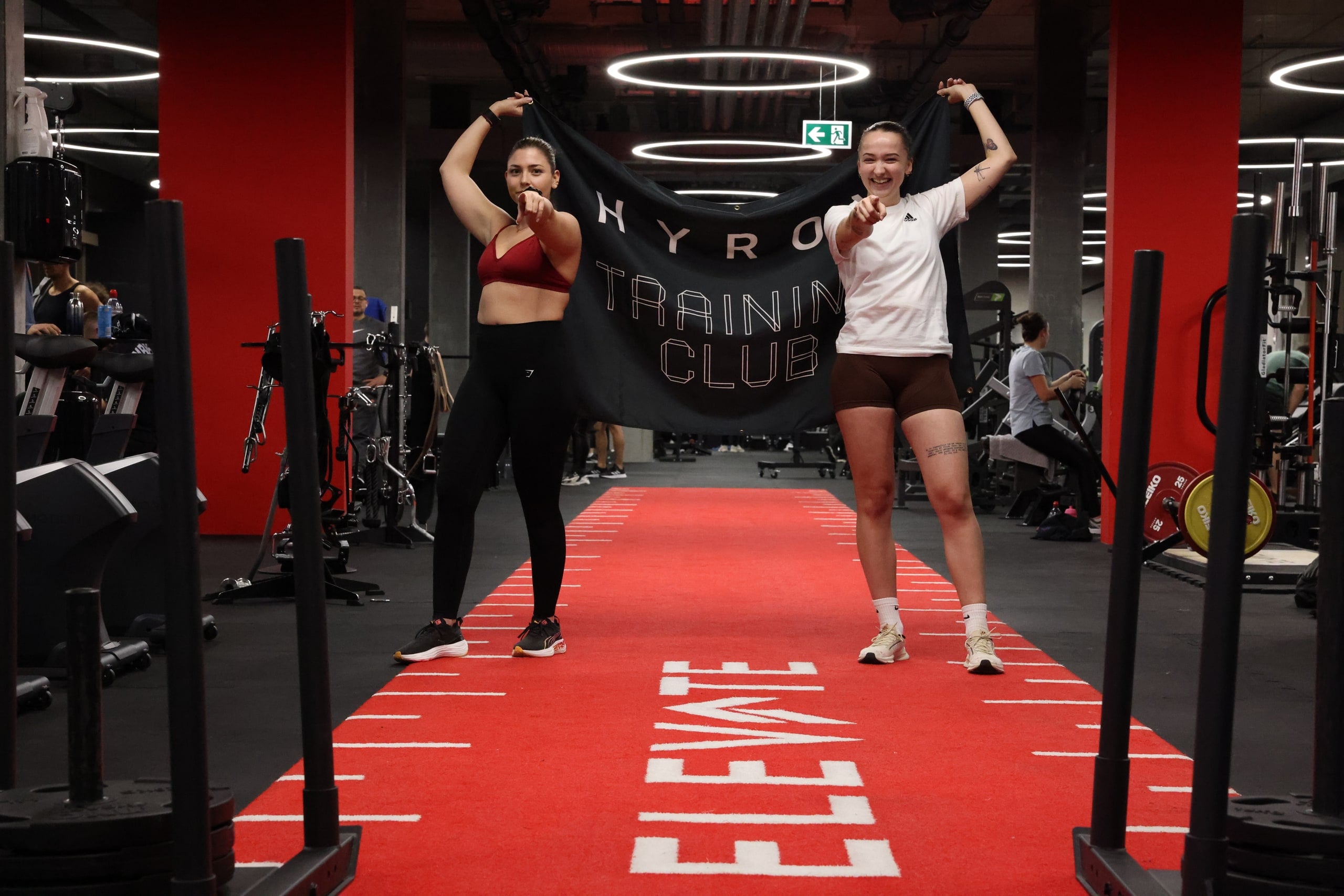 Deux membres du HYROX Training Club brandissent la bannière sur la piste rouge, Women's Day ELEVATE
